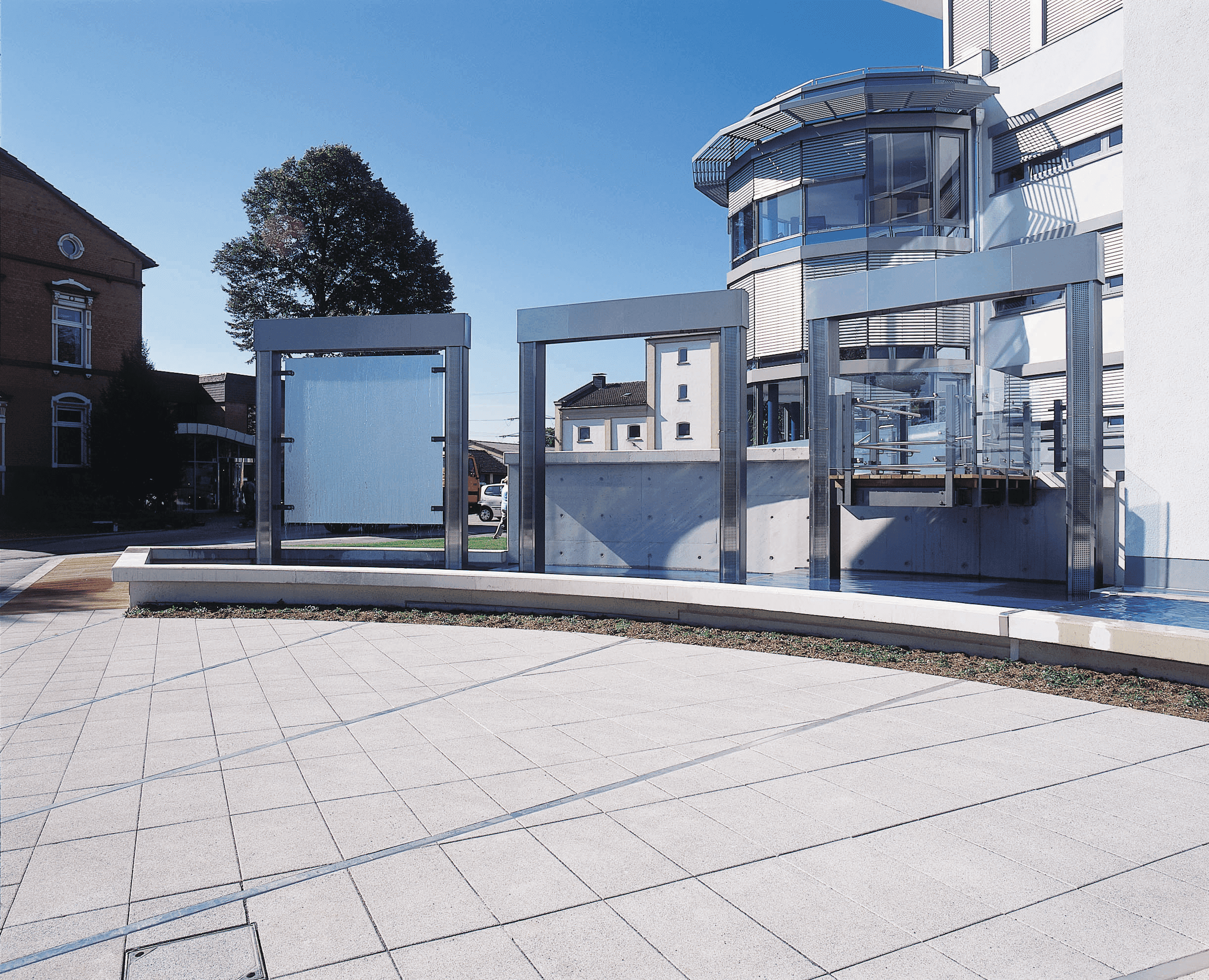 Modern building belonging to Schwerte municipal utilities with glass and metal facade, in front of which are three large glass frames in the outdoor area. To the left is an older brick building. Above is the ‘Stadtwerke Schwerte’ logo.